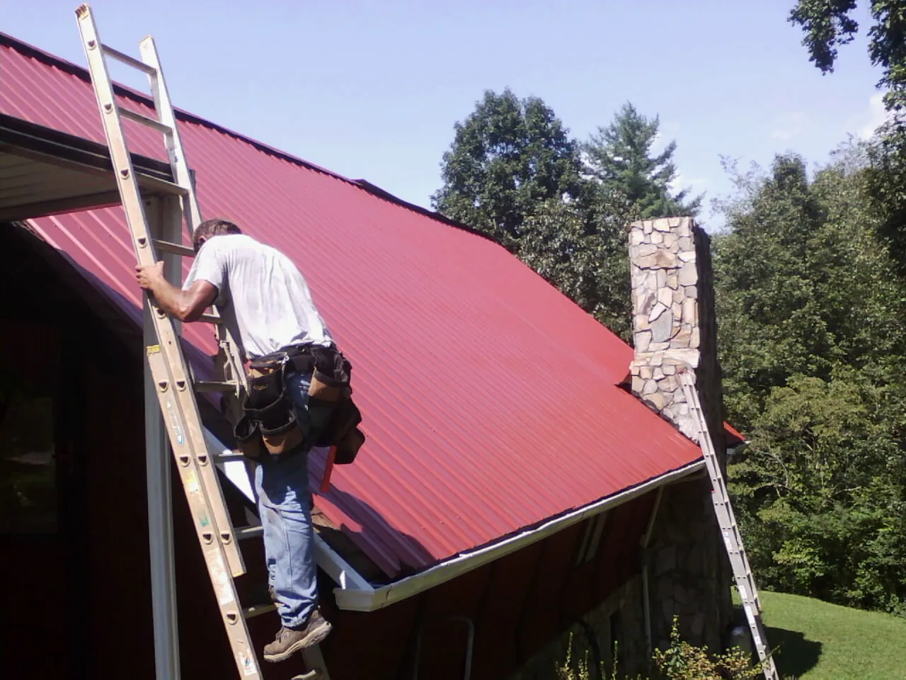 Roofer on a ladder beside a red metal roof and stone chimney — Soffit & Fascia Repair by Roofing Brothers in Stoutsville